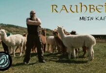 Rauhbein veröffentlichen neue Single „Mein Kaff“ Album cover: bearded man in black with arms crossed among alpacas in a sunny pasture, with 'Raubbein' and 'Mein Kaff' at the top.