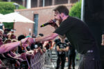 Male singer in a black t-shirt leans toward the crowd, shaking hands with fans during an outdoor concert.