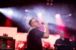 Male singer wearing glasses performs on stage with a handheld mic; keyboardist in the background under pink-red lights.