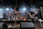 Rock band on stage: singer jumps mid-song, guitarist on the right, drummer center, bright spotlights and a banner behind.