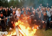 Three Days Grace veröffentlichen Musikvideo zum Song „Don’t Wanna Go Home Tonight“ Crowd of friends around a large bonfire while three musicians play guitars outdoors at dusk, Vevo logo bottom lefting on screen.