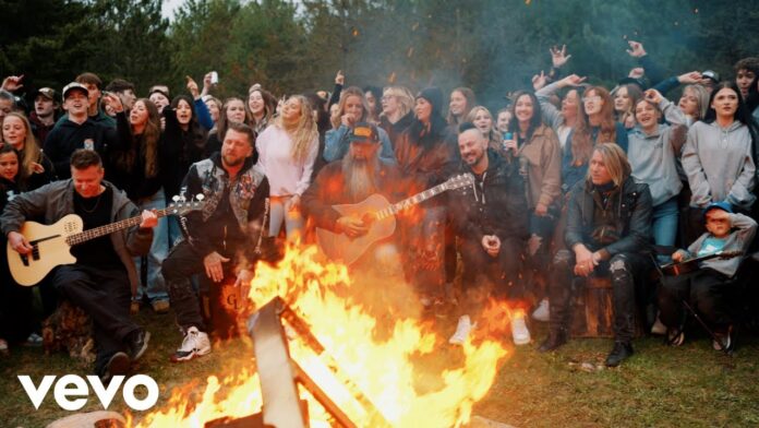 Three Days Grace veröffentlichen Musikvideo zum Song „Don’t Wanna Go Home Tonight“ Crowd of friends around a large bonfire while three musicians play guitars outdoors at dusk, Vevo logo bottom lefting on screen.
