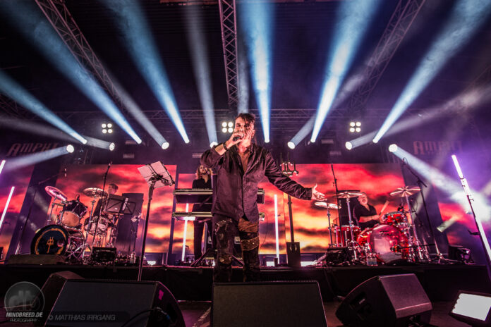 Das war das Amphi Festival 2025 Male lead singer with microphone center stage, band performing with drums and keyboard under bright concert lights.
