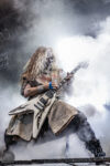 Guitarist in medieval-style costume with skull makeup playing a white flying-V guitar amid stage fog.