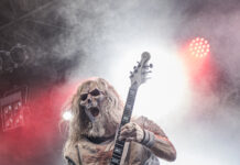 Warkings und Gitarrist „Crusader“ gehen getrennte Wege Guitarist wearing a skull mask and tattered costume plays a white Flying V guitar on a smoky stage with red lights.