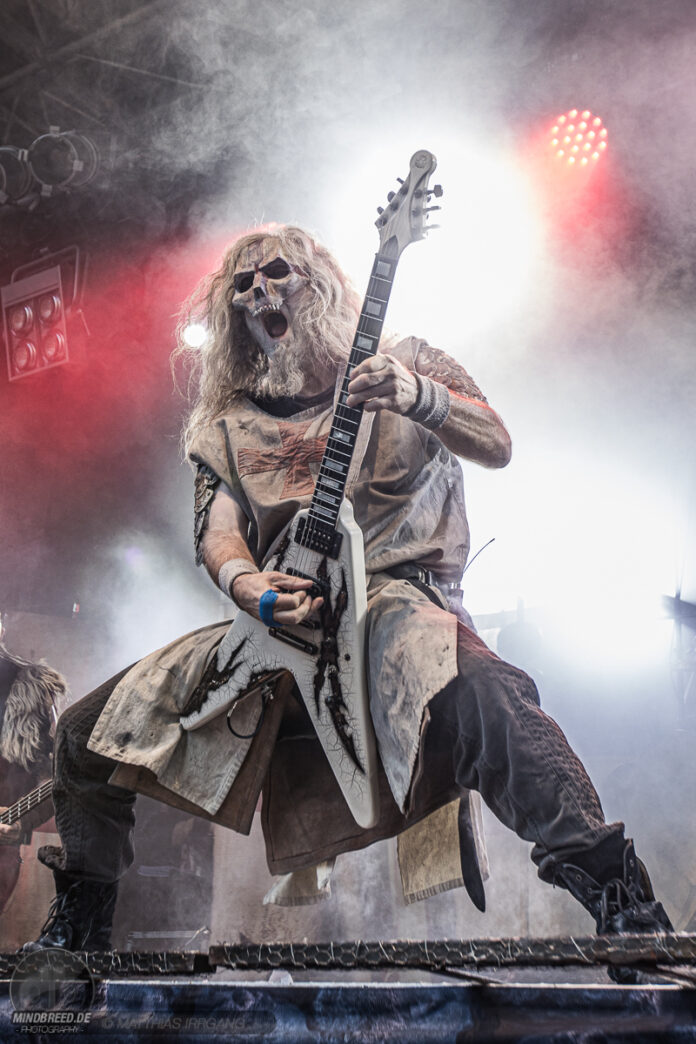 Warkings und Gitarrist „Crusader“ gehen getrennte Wege Guitarist wearing a skull mask and tattered costume plays a white Flying V guitar on a smoky stage with red lights.