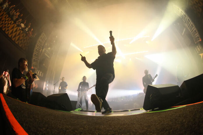 Dropkick Murphys 24.07.26, Store Vega, Kopenhagen Male lead singer on a bright concert stage, mic raised, striking a dynamic pose toward the crowd with band members and lights in the background