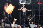 Male vocalist with arms raised on stage as a guitarist blasts fire from his mouth, performing intensely on a metal concert stage.