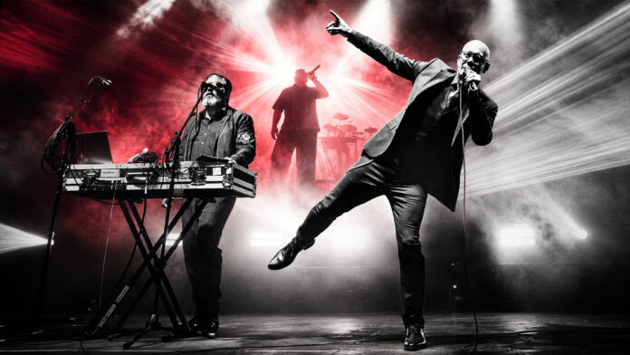Amphi Festival 2026: Covenant springen für VNV Nation ein Band on stage: singer with microphone to the right, keyboardist on the left, drummer in back, dramatic red and white lights filling the scene.