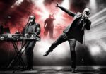 Live concert scene: keyboardist on left and lead singer mid-song on the right, with dramatic red and white stage lights and smoke behind them.