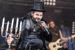 Gothic rock singer in a top hat and black velvet outfit performs on stage, gripping a microphone beside a silver candelabrum.