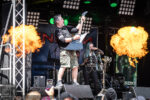 Guitarist in camouflage shorts and black shirt plays a V-shaped guitar on a smoky stage with large flame projections on both sides while a female singer performs nearby.