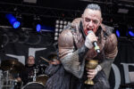 Metal vocalist in rusted armor and chainmail screams into a microphone on stage, holding a golden chalice; drummer in background.