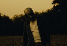 Converge veröffentlichen Video zu „It Used to Matter“ – kündigen EU/UK-Tour an Person with a wrapped head (bandages) and white shirt under a dark jacket stands in a tall grass field at dusk, looking down.