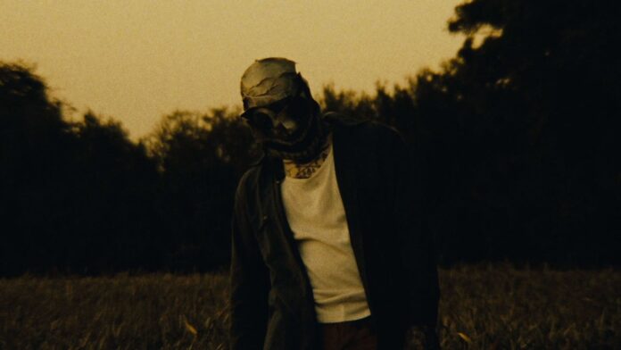 Converge veröffentlichen Video zu „It Used to Matter“ – kündigen EU/UK-Tour an Person with a wrapped head (bandages) and white shirt under a dark jacket stands in a tall grass field at dusk, looking down.