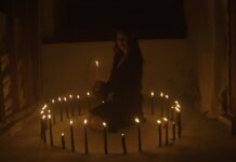 Chelsea Wolfe veröffentlicht neue Single und kündigt Album an Woman sitting cross-legged inside a ring of lit candles in a dark, unfinished room, holding a candle.