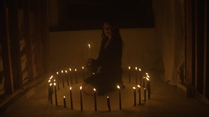 Chelsea Wolfe veröffentlicht neue Single und kündigt Album an Woman sitting cross-legged inside a ring of lit candles in a dark, unfinished room, holding a candle.