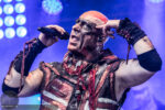 Male rock/metal singer on stage, mouth open mid-song, wearing red headband and dark makeup, gripping a microphone with a distressed costume and bandages under blue stage lights.