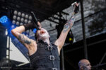 Male rock singer on stage, white face paint, black sleeveless outfit, microphone in hand, arm raised in performance amid blue stage lights and tattoos on both arms.