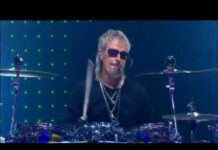 ZZ-Top-Schlagzeuger Frank Beard ist im Alter von 77 Jahren gestorben Drummer with gray hair and sunglasses playing a drum kit on stage, lit by blue lights and green LED backdrop.