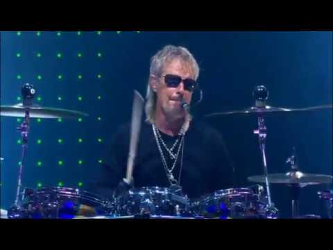 ZZ-Top-Schlagzeuger Frank Beard ist im Alter von 77 Jahren gestorben Drummer with gray hair and sunglasses playing a drum kit on stage, lit by blue lights and green LED backdrop.