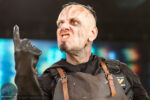 Performer in horror makeup points upward during a stage act, wearing a black shirt and brown leather apron.