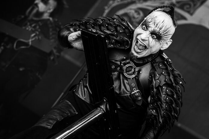 Das Ich: Bruno Kramm nach Konzert in Granada verstorben Black-and-white photo of a performer in spiked leather armor shouting while gripping a large prop weapon on stage.