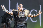 Male performer on stage with black tribal face paint, wearing leather armor and faux bone necklace, arms raised, microphone in hand