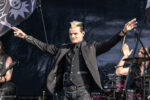 Male rock vocalist on stage with arms wide open, wearing black coat and vest, performing with a band.