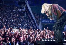 Linkin Park veröffentlichen das offizielle Video zu „Faint“ Singer with blond hair leans into the crowd on a lit concert stage, microphone in hand, surrounded by excited fans taking photos and cheering.