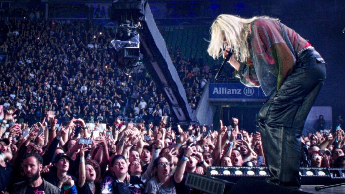 Linkin Park veröffentlichen das offizielle Video zu „Faint“ Singer with blond hair leans into the crowd on a lit concert stage, microphone in hand, surrounded by excited fans taking photos and cheering.