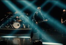 Nickelback präsentiert Musikvideo „Leave Me Behind“ Rock band performing on a dimly lit stage; guitarist center with microphone, drummer left, bassist right under blue beams of light
