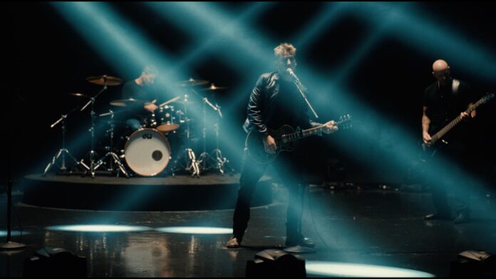 Nickelback präsentiert Musikvideo „Leave Me Behind“ Rock band performing on a dimly lit stage; guitarist center with microphone, drummer left, bassist right under blue beams of light