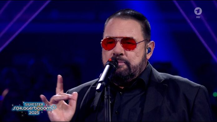 Alphaville werden aus- und wiedereingeladen beim Glücksgefühle Festival Male singer in a black suit with red sunglasses performs on stage into a microphone, blue-lit background visible.