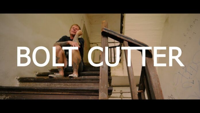 Evergreen Terrace veröffentlichen nächsten Monat eine Coverversion von Jimmy Eat Worlds „Bleed American“ Thumbnail for a video titled 'BOLT CUTTER' showing a man seated on a dusty, decayed staircase in an abandoned building with graffiti in the background.