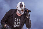 Singer with white and black clown makeup gripping a microphone and singing on stage in a black blazer.