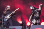Two performers on stage: a guitarist with long hair and white makeup, and a dancer in black lace with a horned headpiece, under red lights.