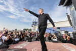 Male singer in black shirt on stage pointing toward the crowd at an outdoor concert, photographers in the foreground.