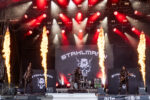Heavy metal band performs on a lit stage with three guitarists and a central vocalist as flames shoot upward from the stage and a skull-bannered backdrop looms behind them.
