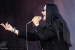 Rock singer with long black hair, sunglasses, and a black jacket passionately singing into a microphone with an outstretched hand on stage.