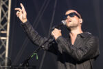 Male singer in sunglasses wearing a dark shirt, singing into a microphone on stage with one hand raised.