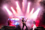 Rock band performs on stage with guitarist center and female vocalist on the right under pink and white lights.