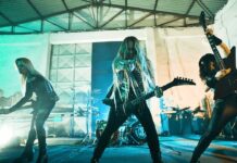Nervosa sagen Termine in UK wegen gesundheitlichen Problemen ab Three female rock musicians perform on a stage in a warehouse, center guitarist with long blond hair exploding energy toward the audience, others on either side playing guitars.