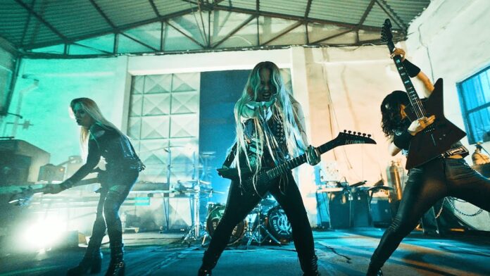 Nervosa sagen Termine in UK wegen gesundheitlichen Problemen ab Three female rock musicians perform on a stage in a warehouse, center guitarist with long blond hair exploding energy toward the audience, others on either side playing guitars.