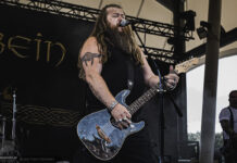 Rauhbein starten im Oktober 2026 auf „Rebellen & Helden“-Tour Bearded male guitarist sings into a microphone while playing a blue guitar on stage, in a black sleeveless shirt and wristbands.