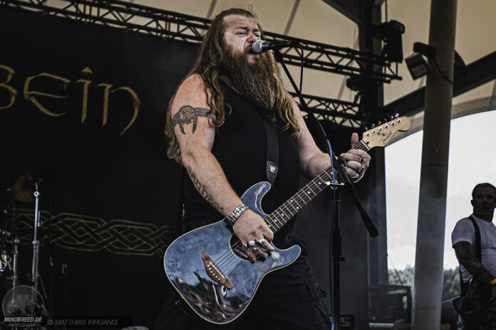 Rauhbein starten im Oktober 2026 auf „Rebellen & Helden“-Tour Bearded male guitarist sings into a microphone while playing a blue guitar on stage, in a black sleeveless shirt and wristbands.